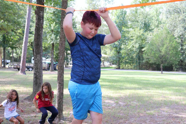 A young boy dressed in blue holds onto an amber colored line above his head as he practices walking tightrope at summer camp