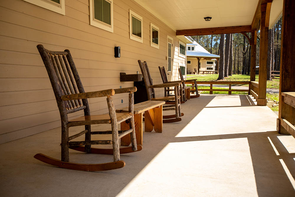 Four live-edge wooden rocking chairs sit facing outward to the right on a sunny porch retreat with tables between each set of chairs in groups of two.