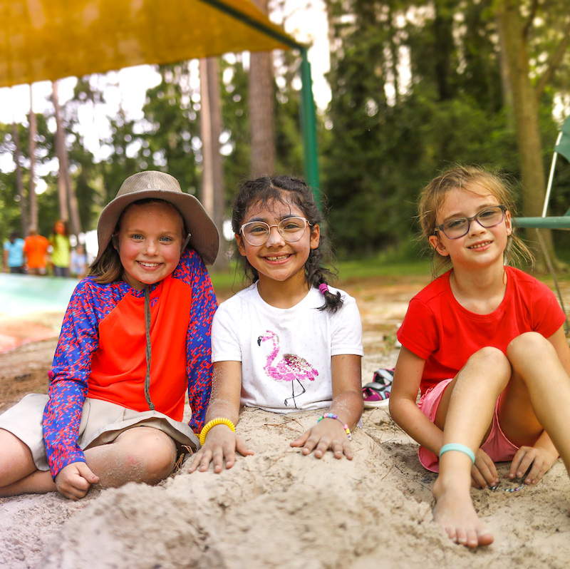 Three young girls sit in a pile of sand grinning at the camera while at summer camp. Two of the girls have on glasses and another has a hat.